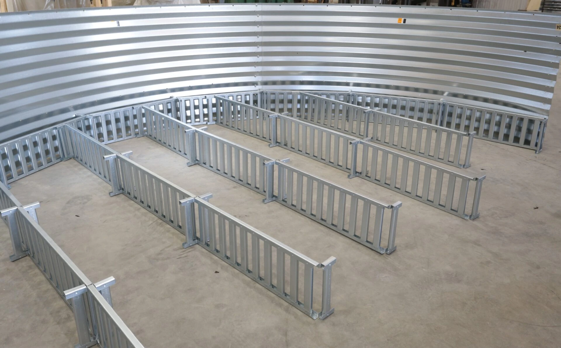 A curved, metallic structure with vertical slatted dividers arranged on a concrete floor, likely a livestock confinement pen